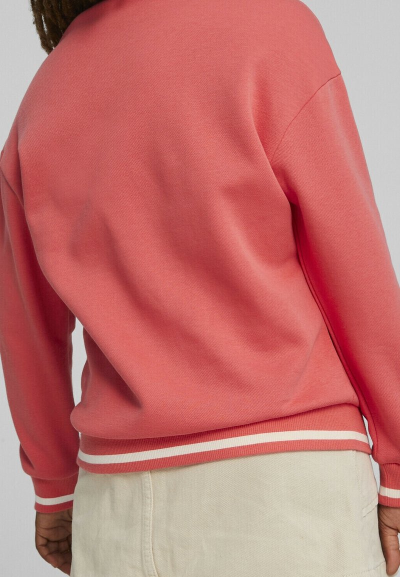 Sweatshirt corail avec des poignets et un ourlet côtelés, comportant des rayures blanches en accent. Texture douce avec une coupe décontractée. Vue du dos et détails de la couture latérale.
