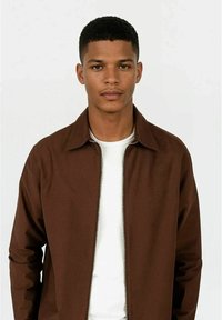 Brun zip-up jakke med krage, laget av et glatt materiale, brukt over en hvit t-skjorte. Har et enkelt design og synlig glidelås.