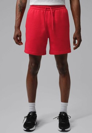 Hombre con pantalones cortos rojo brillante, calcetines blancos y zapatos deportivos negros, de pie frente a un fondo gris liso.