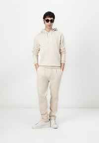 Felpa in cotone beige con coulisse, texture a fantasia, abbinata a pantaloni jogger coordinati. Sneakers bianche completano l'outfit.