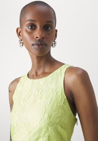 Robe sans manches vert lime avec une texture florale en relief ; décolleté rond ; mannequin portant des boucles d'oreilles en argent ; teint de peau lisse.