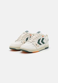 White sneakers featuring green accents, textured leather, round toe, and orange outsole details. Laces and logo on tongue visible.