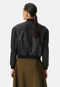Giubbotto bomber in pelle nera con polsini e orlo a coste, caratterizzato da una texture liscia e un design accorciato, indossato sopra una gonna verde oliva.
