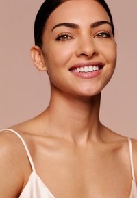 Lächende Person mit braunen Augen und dunklem Haar, die ein helles Satinoberteil trägt. Reine Haut, minimaler Schmuck, neutraler Hintergrund.