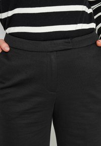 Zwarte broek met een gladde textuur, voorzien van een platte tailleband en een zijzak. De stof lijkt rekbaar en is op maat gemaakt voor comfort.