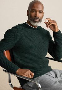 Dunkelgrüner, gerippter Pullover mit Rundhalsausschnitt, aus einem strukturierten Strickstoff gefertigt. Modell sitzt auf einem Stuhl und trägt graue Hosen.