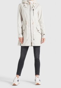 Parka zip in beige chiaro con cappuccio regolabile e cordino in vita, due tasche frontali e una tasca con zip sul petto. Abbinato a jeans neri e sneakers.