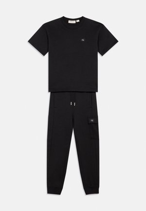 Ensemble t-shirt manches courtes noir et pantalon jogger avec petites écussons logo sur la poitrine et la poche de la cuisse, présenté sur fond blanc.