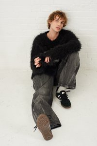 Black fluffy sweater, gray baggy jeans, and black sneakers with white accents. Natural hair styled in loose curls, seated against a white wall.