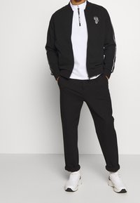 Giacca bomber nera con zip e dettagli bianchi, abbinata a una maglietta bianca e pantaloni neri con maniche arrotolate e sneaker bianche.