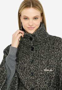 Cappotto grigio a motivo houndstooth con colletto alto, bottoni neri e maniche rigate grigie. Presenta un logo ricamato sul petto.