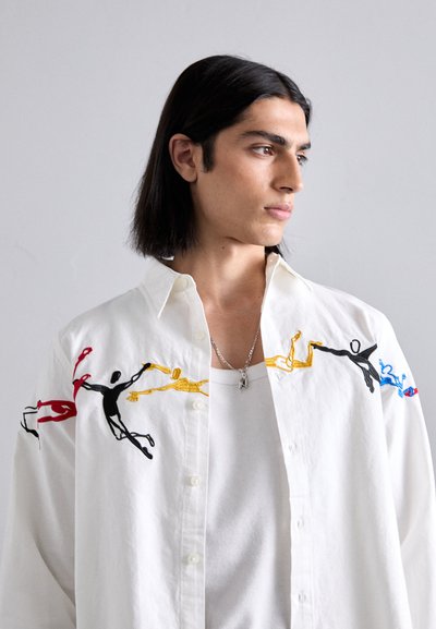 White button-up shirt with colorful embroidered figures in red, yellow, black, and blue on the sleeves. Smooth fabric texture.