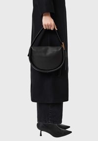 Čierna kožená kabelka s zaobleným tvarom, kontrastným dreveným akcentom a strapcovým detailom, držená rukou s prsteňom.