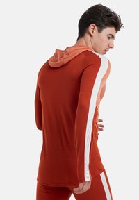 Felpa rossa a maniche lunghe con cappuccio arancione chiaro. Presenta righe bianche sui lati. Texture morbida, design aderente e stile casual adatto per lo sport.
