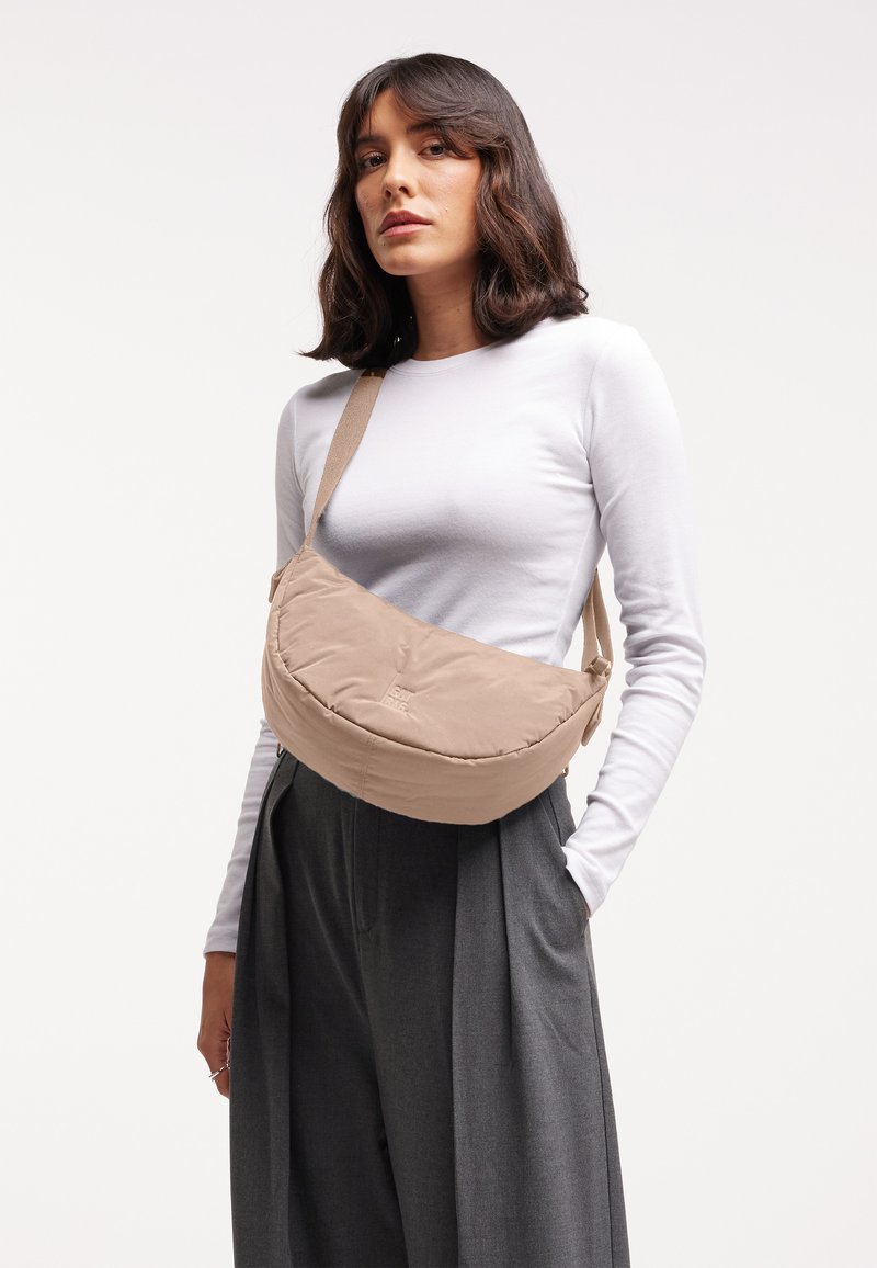 Mujer con cabello oscuro que lleva una camisa blanca de manga larga, pantalones grises y un bolso bandolera beige sobre un fondo liso.