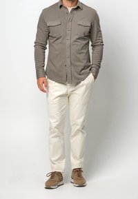 Chemise grise à manches longues et boutonnée avec deux poches poitrine, portée avec un pantalon couleur crème et des baskets en daim marron, sur un fond clair.