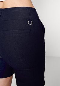 Nærbillede af sorte tætsiddende shorts med baglomme med påsyet kant og lille sølvringdetalje nær linningen på en lys baggrund.