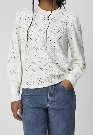 Person iført en hvid langærmet blondebluse med blomsterprint og blå denimjeans, hånd i lommen mod en ensartet baggrund.