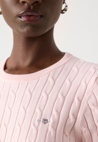 Vaaleanpunainen neulepaita, jossa on kaapelikuvio, ribbireunus kaula-aukossa ja pieni brodeerattu logo rinnassa. Pehmeä rakenne.