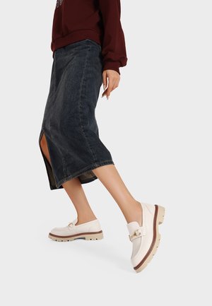 Scarpe slip-on bianche con suola spessa e dettagli in oro, indossate con una gonna in denim scura con spacco frontale.