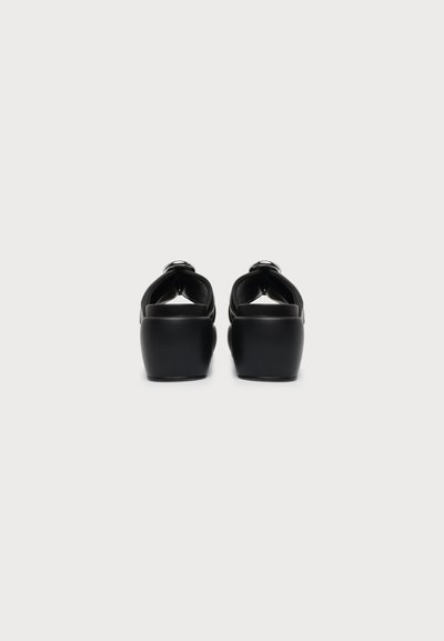 Paire de sandales à plateforme noires avec des talons épais, vues de dos sur un fond clair uni.