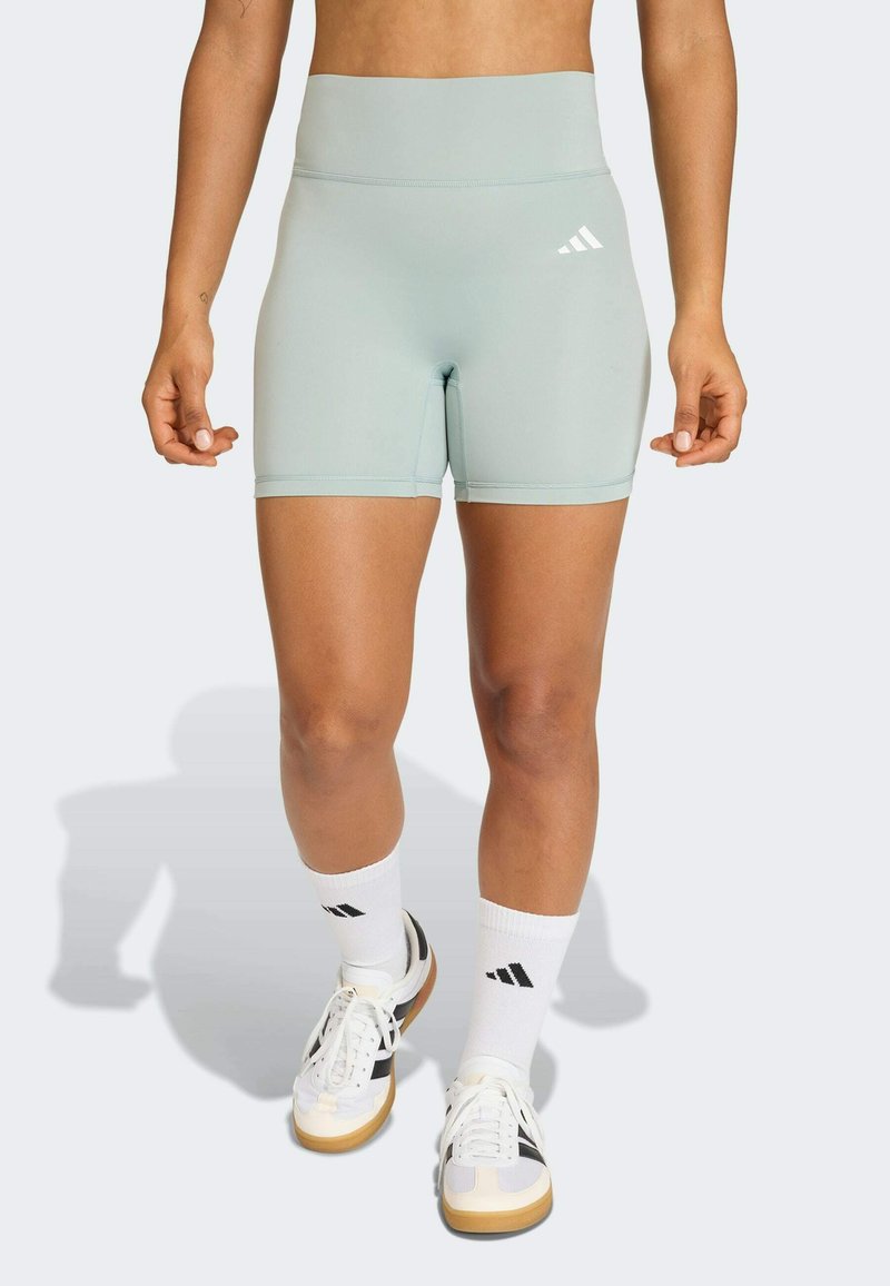 Shorts taille haute vert clair en matière extensible, avec un logo blanc sur la hanche gauche, assortis à des chaussettes et des baskets blanches.