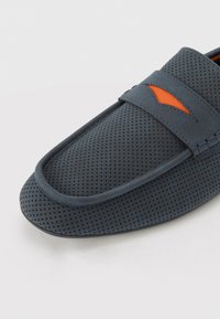 Mørkeblå perforerede slip-on sko med en rund tå og en kontrasterende orange accent synlig over remmen. Tekstureret overdel og glat sål.