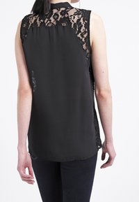 Haut sans manches noir avec un dos en dentelle texturée, un devant lisse, un ourlet arrondi et un éclat subtil dans le tissu.