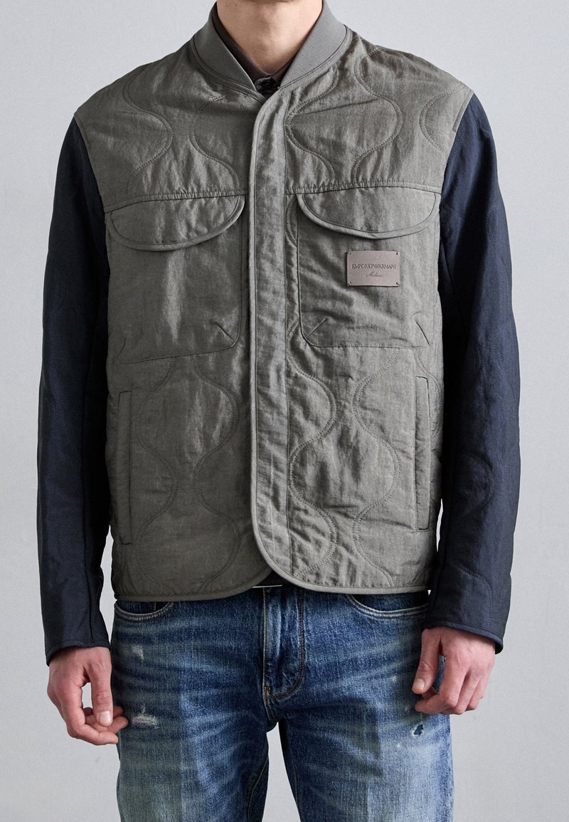 Mężczyzna ubrany w pikowaną szarą kamizelkę z czarnymi rękawami i niebieskie dżinsy, stojący na tle jednolitego jasnoszarego tła.