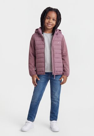 Ung jente med flettehår iført en lilla hettejakke, grå skjorte, blå jeans og hvite joggesko, stående og smilende.