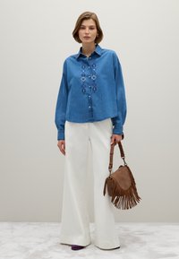 Eine Frau trägt ein blaues besticktes Denim-Hemd, weite weiße Hosen, lila Schuhe und hält eine braune Leder-Handtasche mit Fransen.