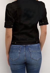 Svart kortärmad jacka med en strukturerad design, som har knapppdetaljer vid ärmarna och fållen, ihop med blå denimjeans med bakfickor.