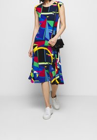 Colorful sleeveless dress featuring a geometric pattern in blue, green, red, yellow, and black; paired with a small black handbag and white sneakers.