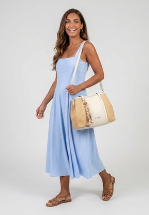 Femme souriante, portant une robe midi sans manches bleu clair, des sandales marron, un sac à bandoulière tissé beige et crème avec un détail de pompon.