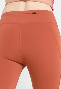 Leggings aderenti a vita alta di colore ruggine, realizzati in materiale elastico. Presentano dettagli delle cuciture e una tasca discreta in vita.
