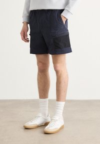 Navy šortky vyrobené z lehkého materiálu, s kapsou na zip, elastickým pasem a kontrastním černým panelem na straně.