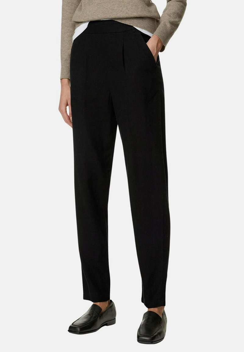 Femme portant un pantalon noir sur mesure, un pull beige et des mocassins noirs, debout avec une main dans la poche devant un fond uni.