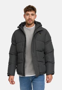 Man in een donkergrijze gewatteerde jas met capuchon, lichtgrijze gebreide trui en blauwe jeans, staande tegen een effen achtergrond.