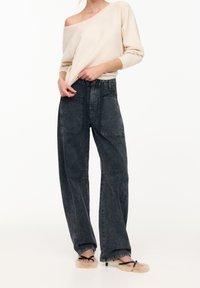 Vaaleahko beige, olkapäilta avattu neulepaita, joka on yhdistetty korkeavyötäröisiin, tummanharmaisiin, leveälahkeisiin farkkuihin, joissa on etutaskut ja haalistunut pinta.