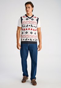 Gebreide mouwloze trui in crème met een feestelijk patroon van rode bomen, zwarte rendieren en sneeuwvlokken. Heeft een V-hals en een geribbelde zoom.