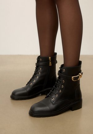 Jambes vêtues de bas portant des bottines en cuir noir à lacets, tirettes de fermeture éclair dorées et sangles décoratives à boucle dorée sur un sol neutre.