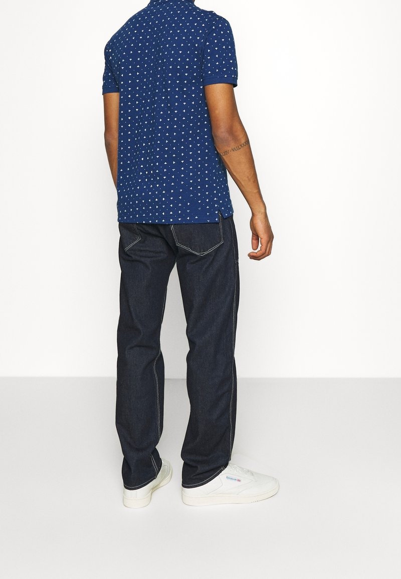 Homme portant un polo bleu à motif, un jean foncé avec des coutures blanches et des baskets blanches, debout sur un fond blanc uni.