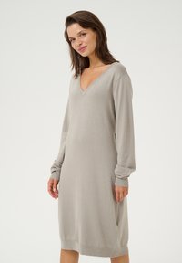 Robe tricotée grise avec des manches longues, un décolleté en V profond et des poignets côtelés. Le tissu a une texture douce et une coupe légèrement ample.