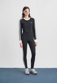 Žena stojí v čiernom teplákovom oblečení Adidas s bielymi pruhmi, v bielych ponožkách, šedých topánkach na suchý zips a bielych športových slnečných okuliaroch na modrej podlahe.