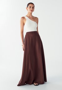 Bruine, vloeiende maxi rok gecombineerd met een witte ribbel, één-schouder top. De rok heeft een gladde textuur en valt elegant tot op de vloer.