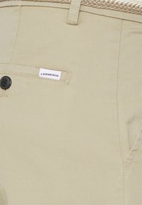 Beige byxor gjorda av vävt material; har en svart knapp, sydda fickdetaljer och en vit etikett med "LINDBERGH" tydligt synlig.