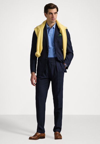 Traje a rayas marinas con un polo azul claro y un suéter de punto amarillo sobre los hombros. Zapatos de vestir marrones. Diseño minimalista.