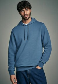 Felpa con cappuccio blu in tessuto testurizzato, con polsini elasticizzati e tasca a marsupio frontale. Abbinata a jeans in denim scuro.