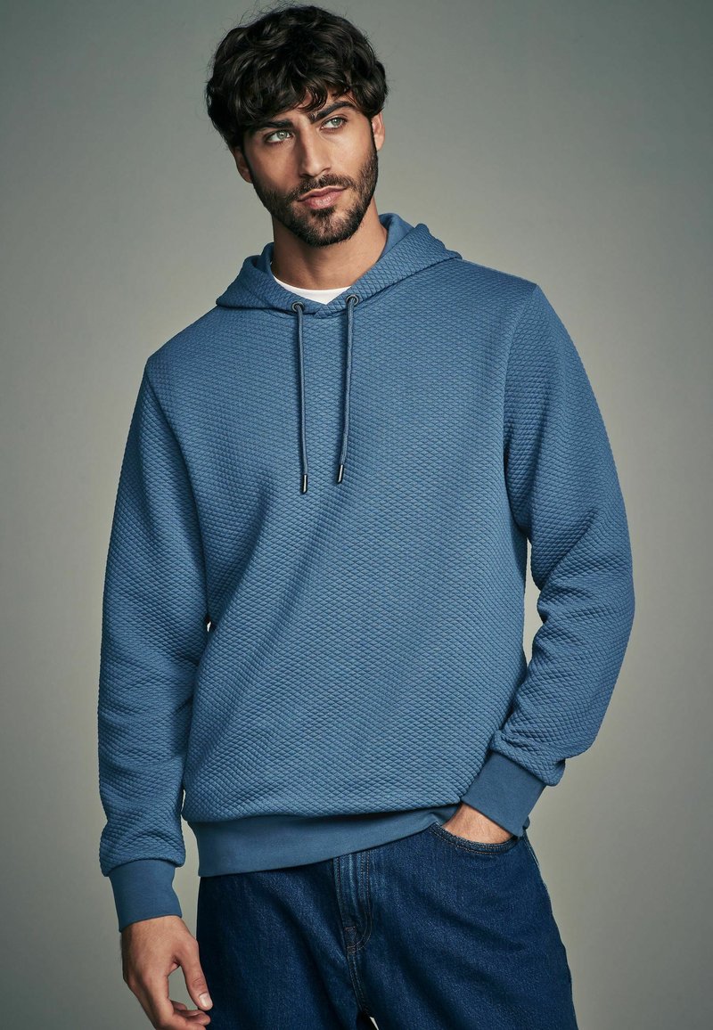 Felpa con cappuccio blu in tessuto testurizzato, con polsini elasticizzati e tasca a marsupio frontale. Abbinata a jeans in denim scuro.