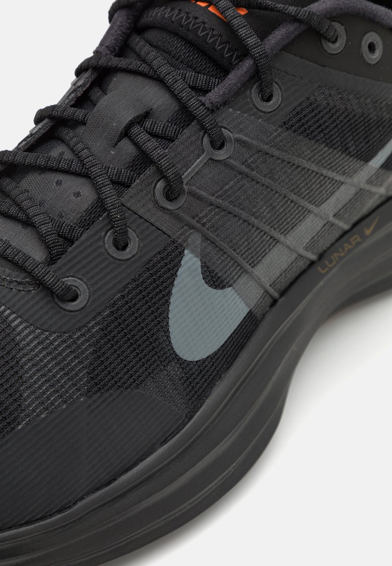 Czarna biega but Nike zbliżenie, pokazujące siatkowy materiał, sznurówki, szary logo swoosh oraz napis "Lunar" na grubej podeszwie na białym tle.